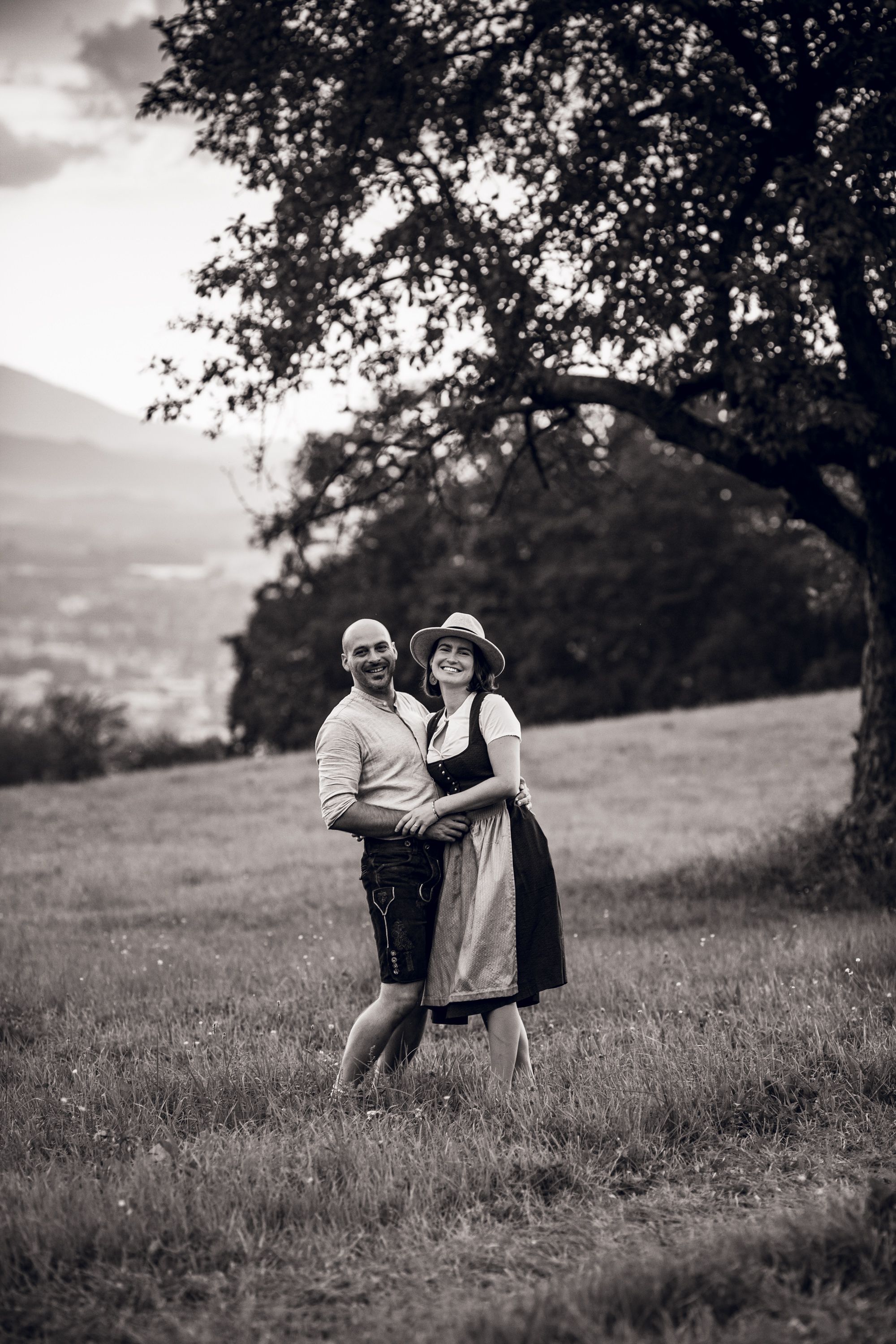 A black and white romantic photograph showing a couple embracing in a scenic rural meadow at sunset.