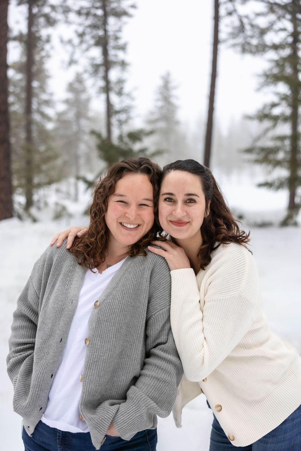 Two people in casual winter wear share genuine laughter against snowy pine trees.