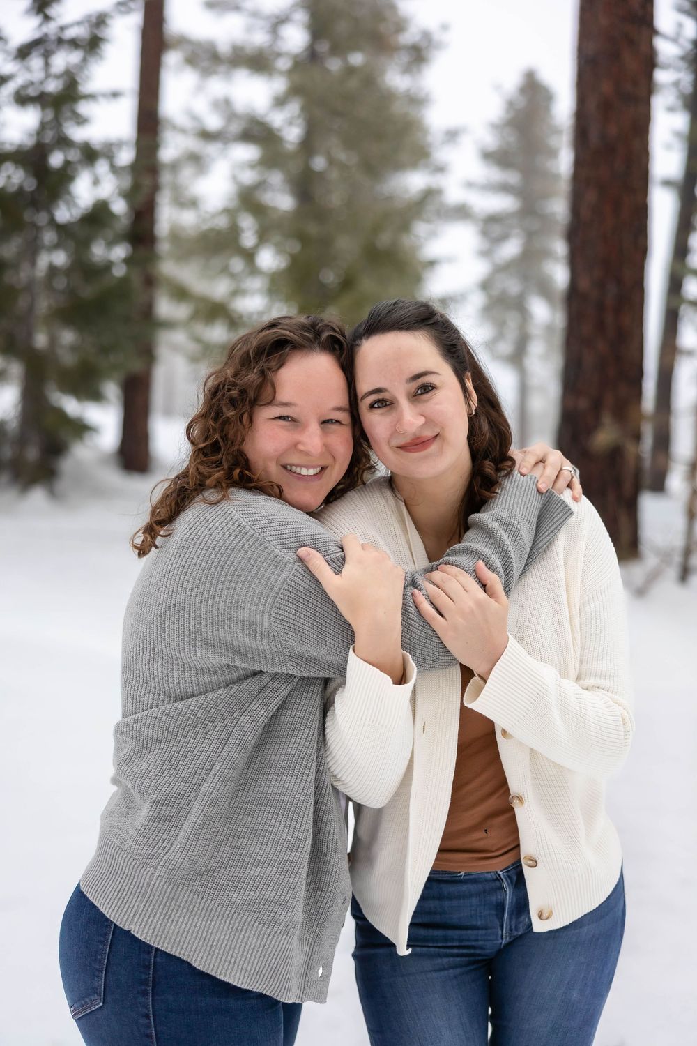 Friends in cozy sweaters laugh and hug in a snow-covered forest setting.