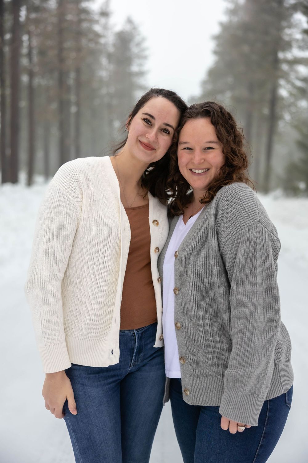 Two people in cardigans share candid moments during a snowy forest photoshoot.