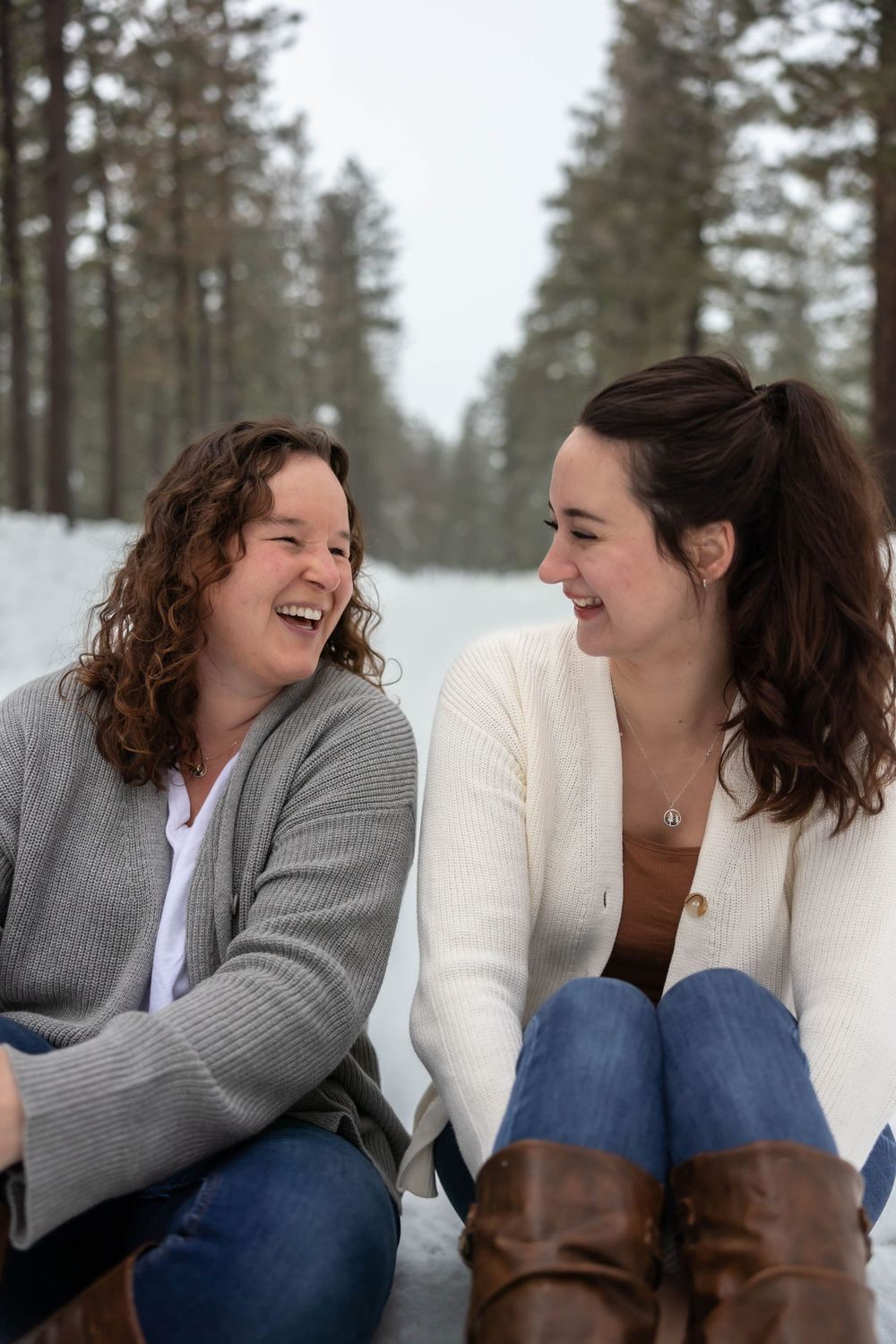 Two people laughing together while sitting outdoors in winter with snow and evergreen trees in the background.
