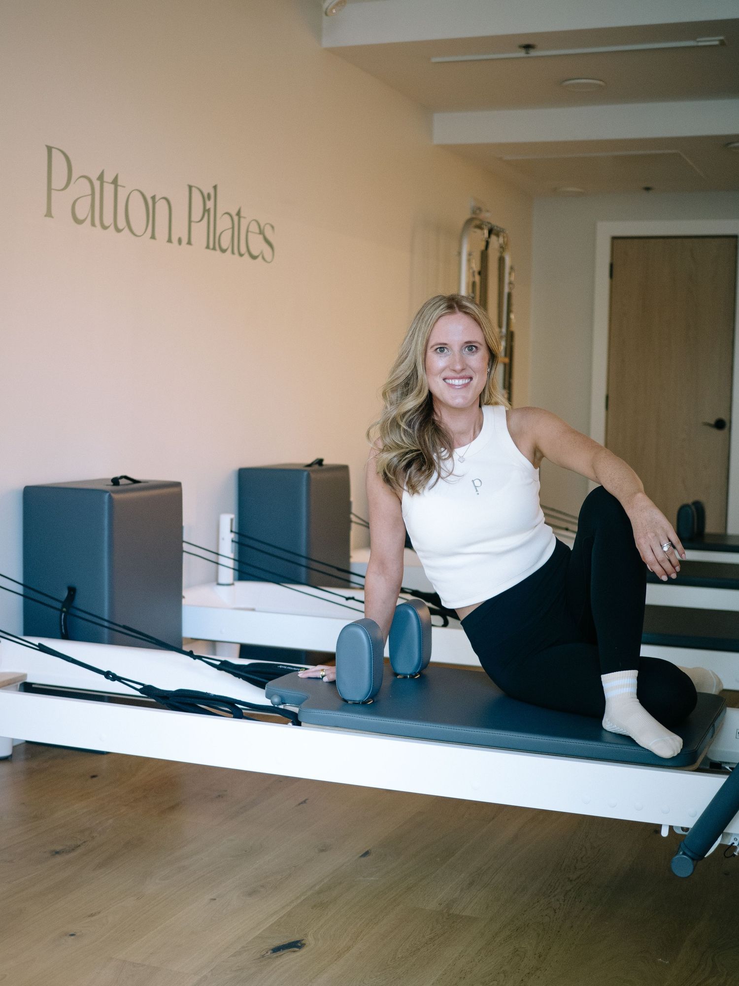 Students practice postures on Pilates reformer machines in a bright studio space with hardwood floors.