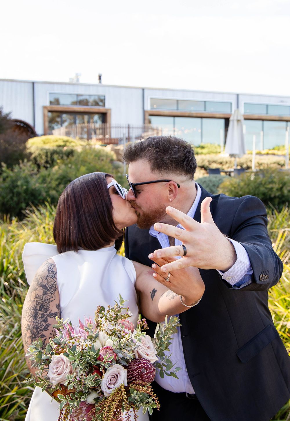 A couple shares a kiss in front of garden foliage while holding a romantic bouquet of pink and burgundy flowers.