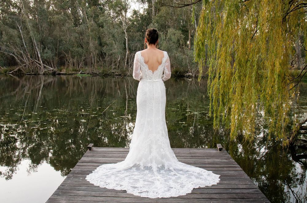 Bride in flowing white lace wedding dress stands on wooden dock by tranquil lake surrounded by willow trees.