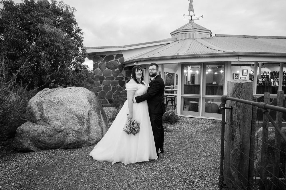 A black and white wedding photo showing a couple embracing outside a building with a large boulder and wooden gate.