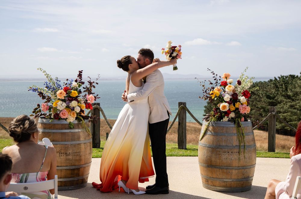 Wedding ceremony on an oceanfront terrace with wine barrels and colorful floral arrangements at sunset.