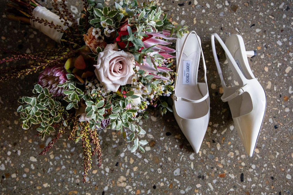 A collection of wedding details including white bridal shoes and floral arrangements laid on a textured stone surface.