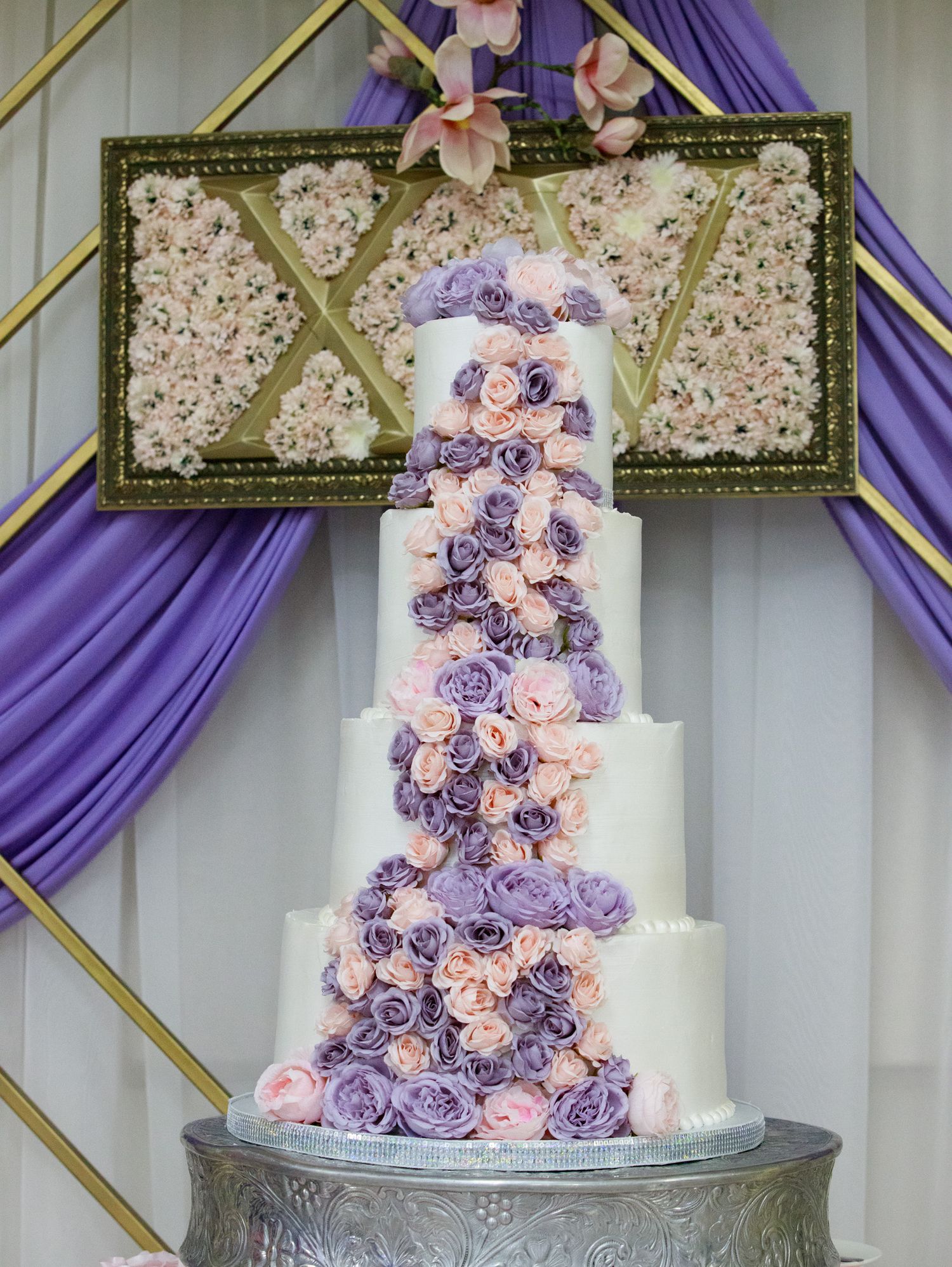 Elegant five-tier white wedding cake decorated with cascading purple and pink roses on a silver stand against draped fabric.