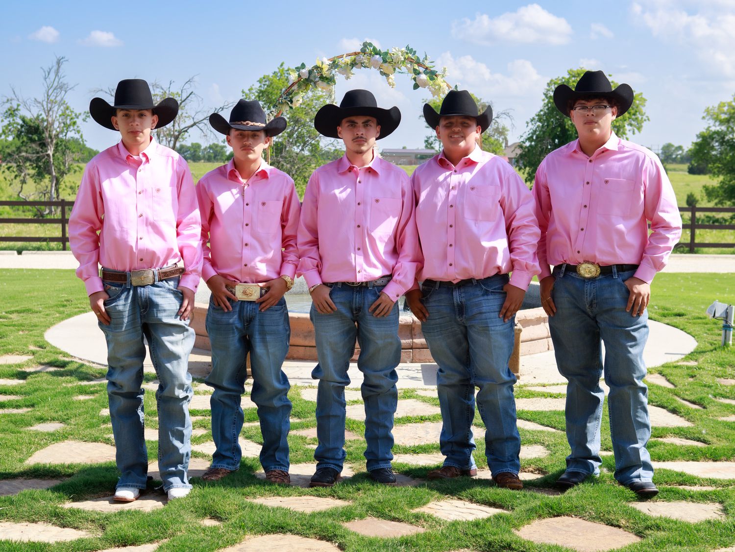 A row of people in western attire with black cowboy hats and pink shirts standing together outdoors on a sunny day.