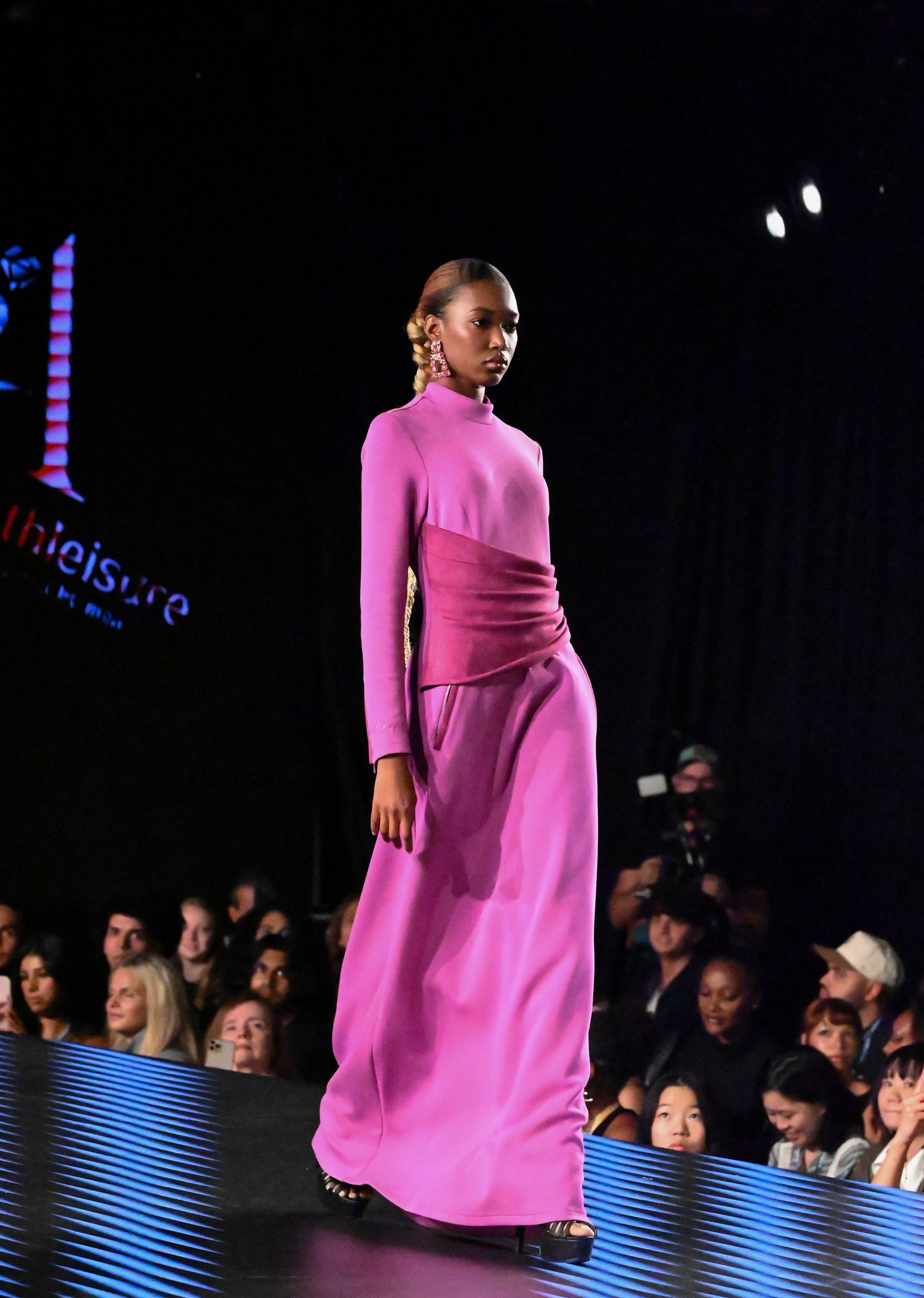 Model walks the runway in vibrant pink high-neck maxi dress during fashion show with audience visible in background.