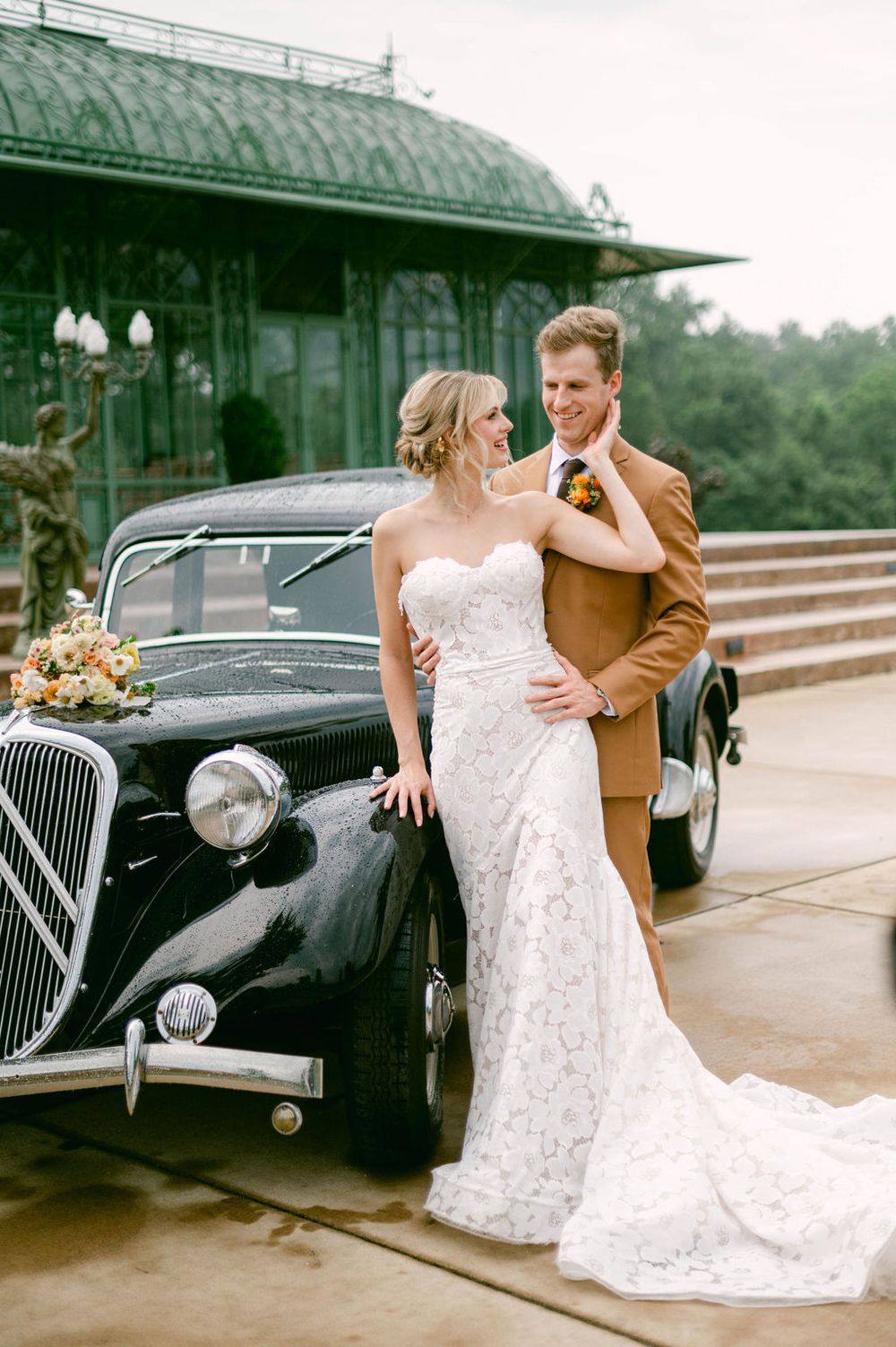 A romantic wedding couple poses with a vintage black car at an old train station.