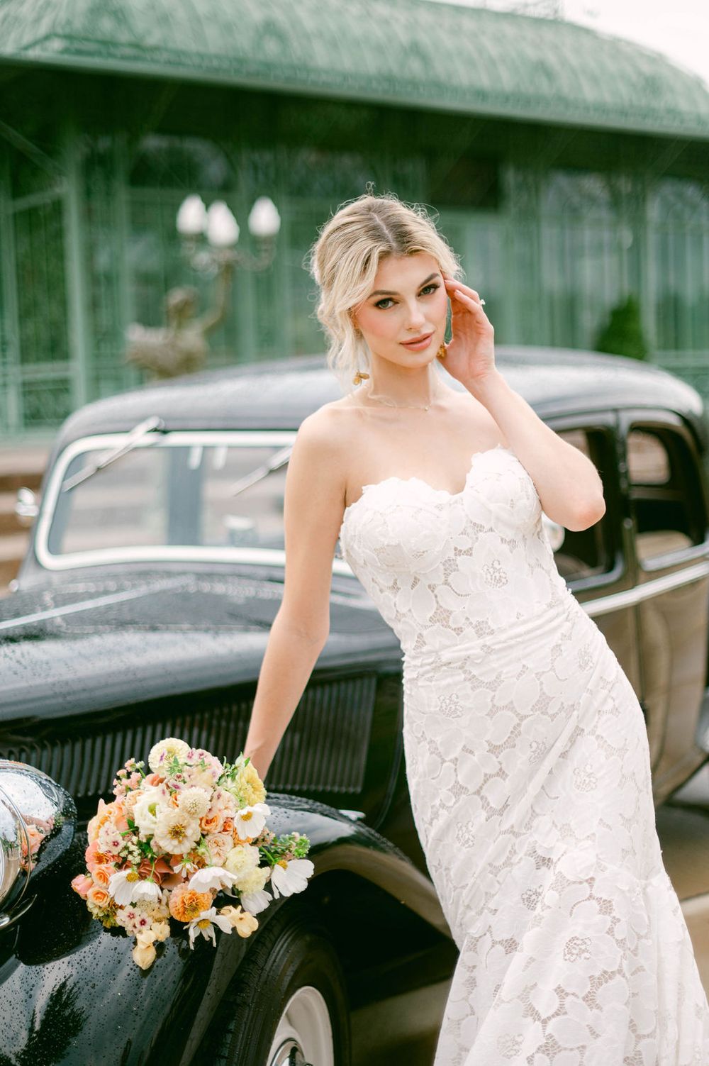 Elegant bride in a white lace dress poses with a peach and cream floral bouquet near a vintage car.