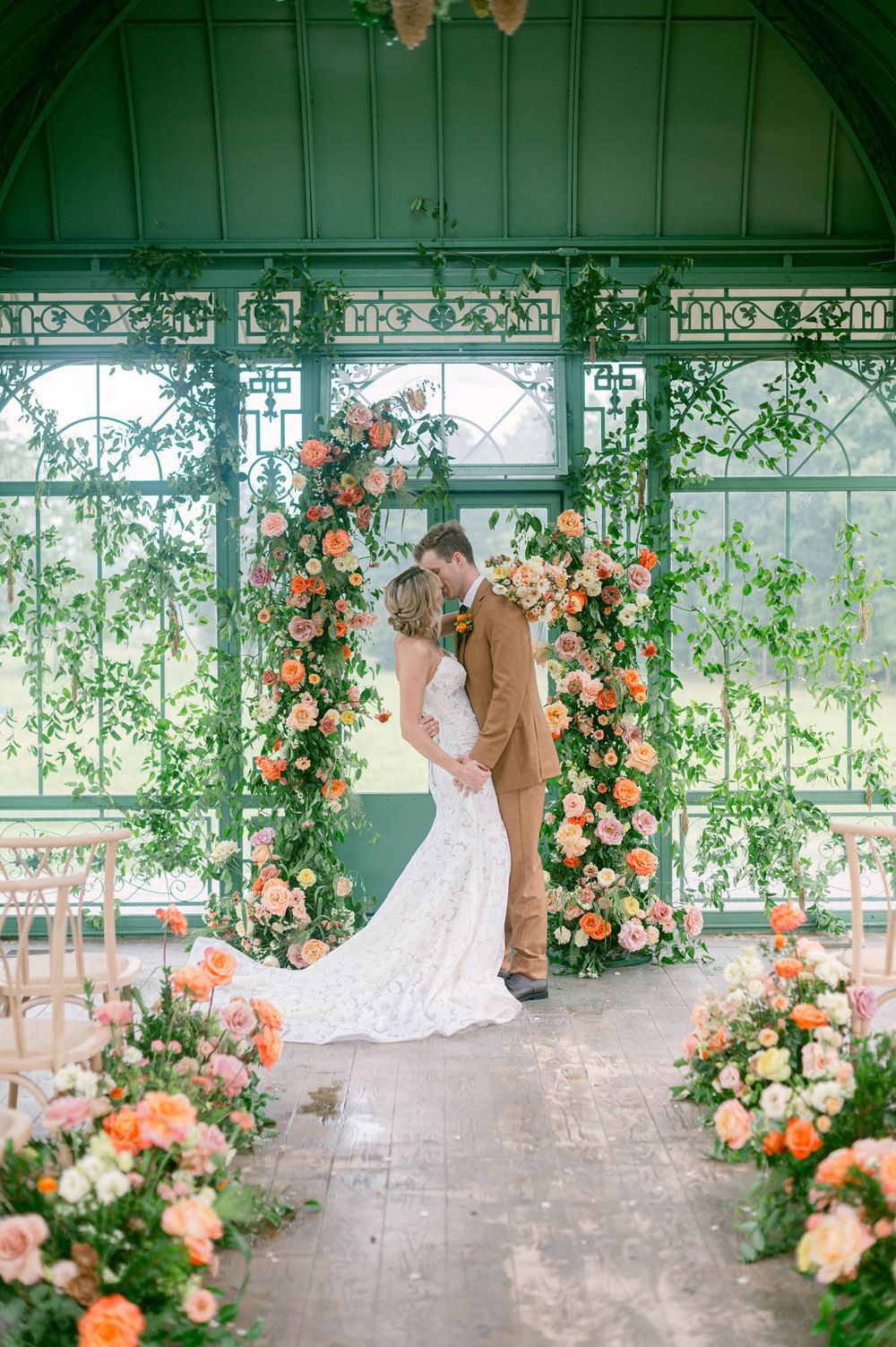 A Morais Vineyard wedding greenhouse ceremony setup features lush greenery and peach floral arrangements along a wooden floor.
