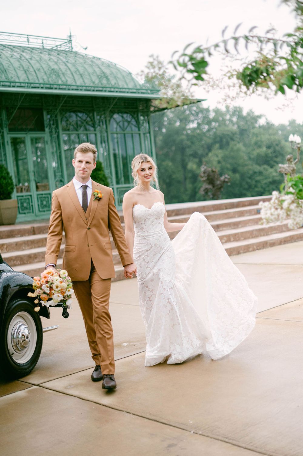 Couple in brown suit and white wedding dress pose together near green vintage structures for outdoor wedding portraits.