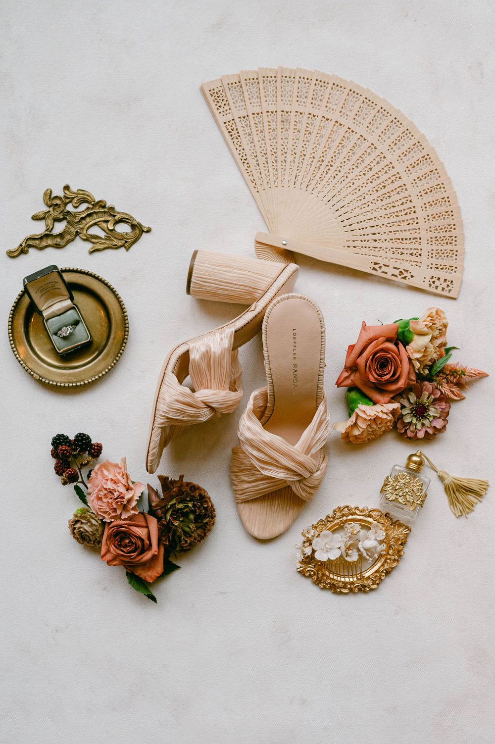 Vintage wedding accessories including pink fans, gold compasses, floral corsages, and blush sandals arranged on white surface.