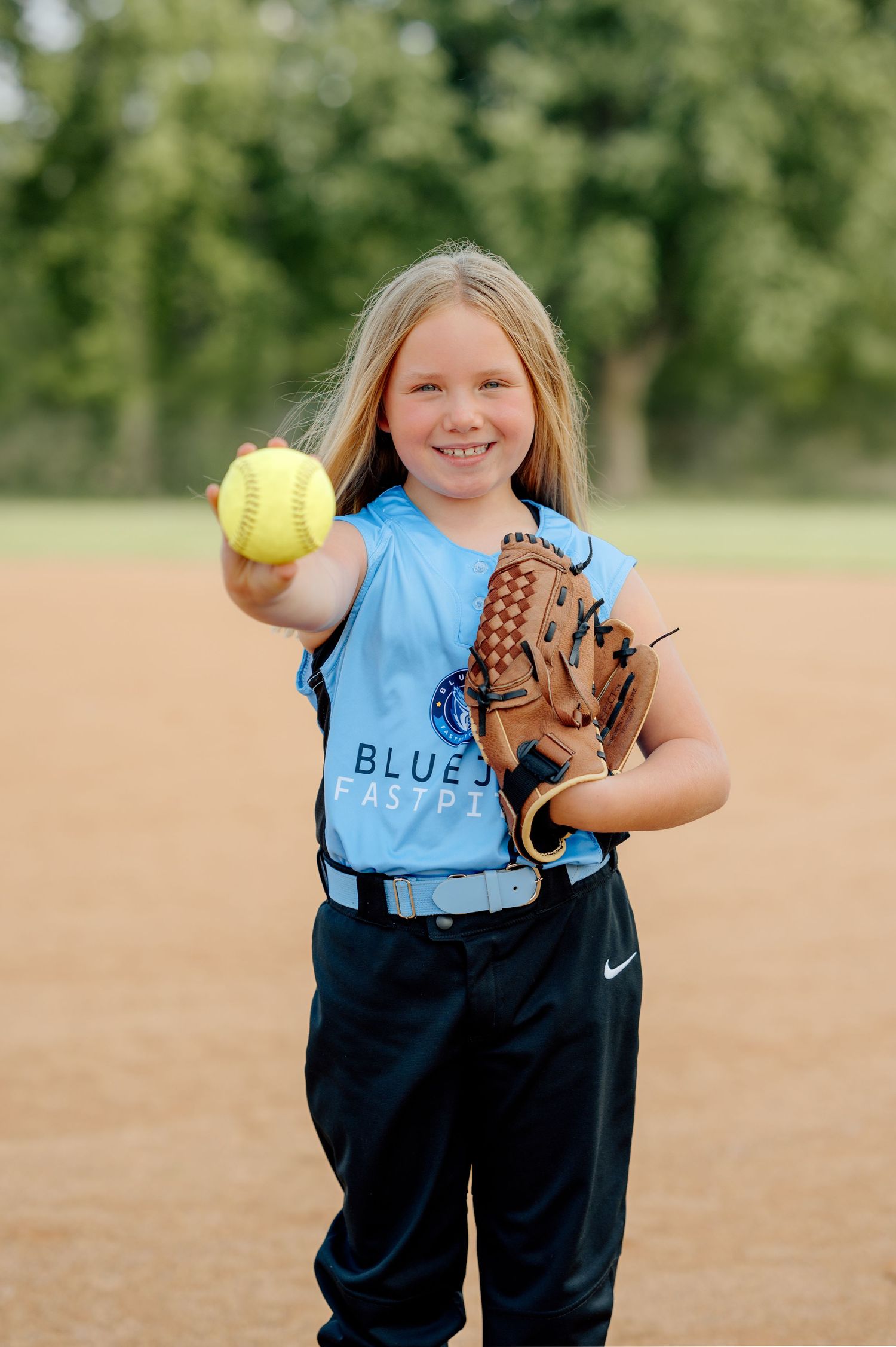 Young softball player in light blue jersey holds up yellow softball while wearing brown leather glove on baseball diamond.