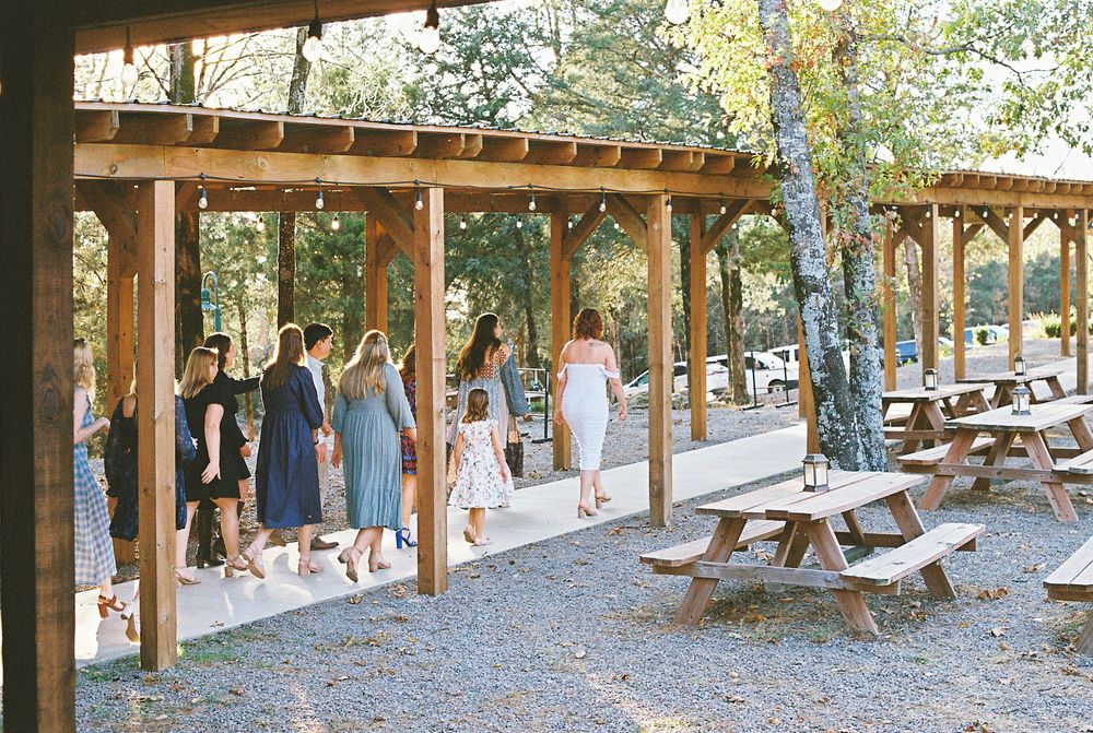 A covered wooden pavilion with picnic tables extends along an outdoor path surrounded by trees in a park setting.