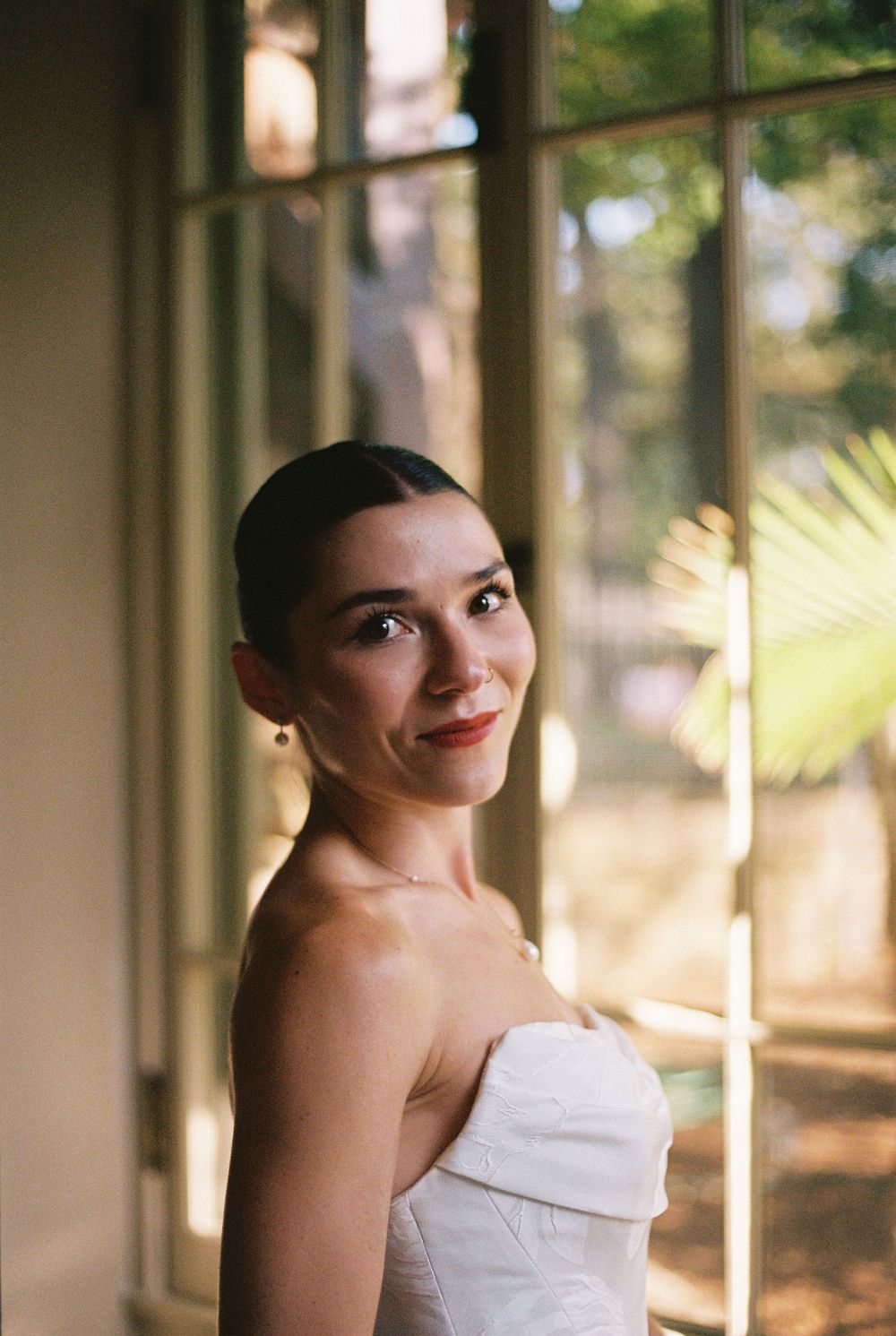 A figure in a strapless white gown poses elegantly by sunlit windows in an indoor setting.