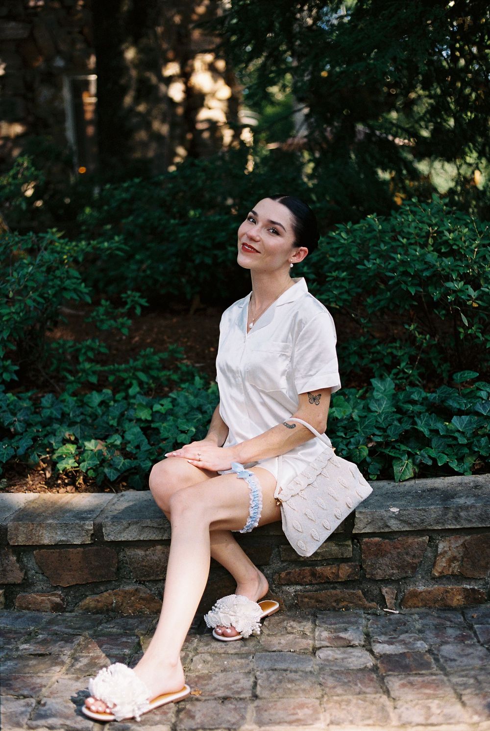 Someone wearing a white top and shorts sits casually on a stone wall surrounded by lush greenery in a garden setting.