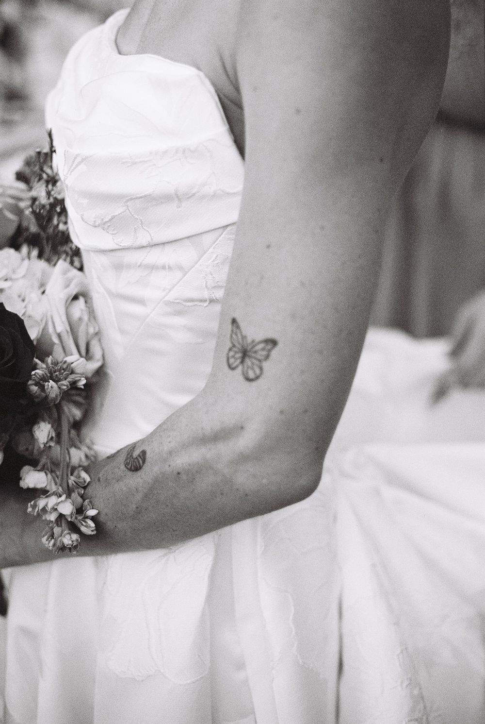 Black and white photo showing a delicate butterfly tattoo on an arm holding a bouquet in a white formal dress.