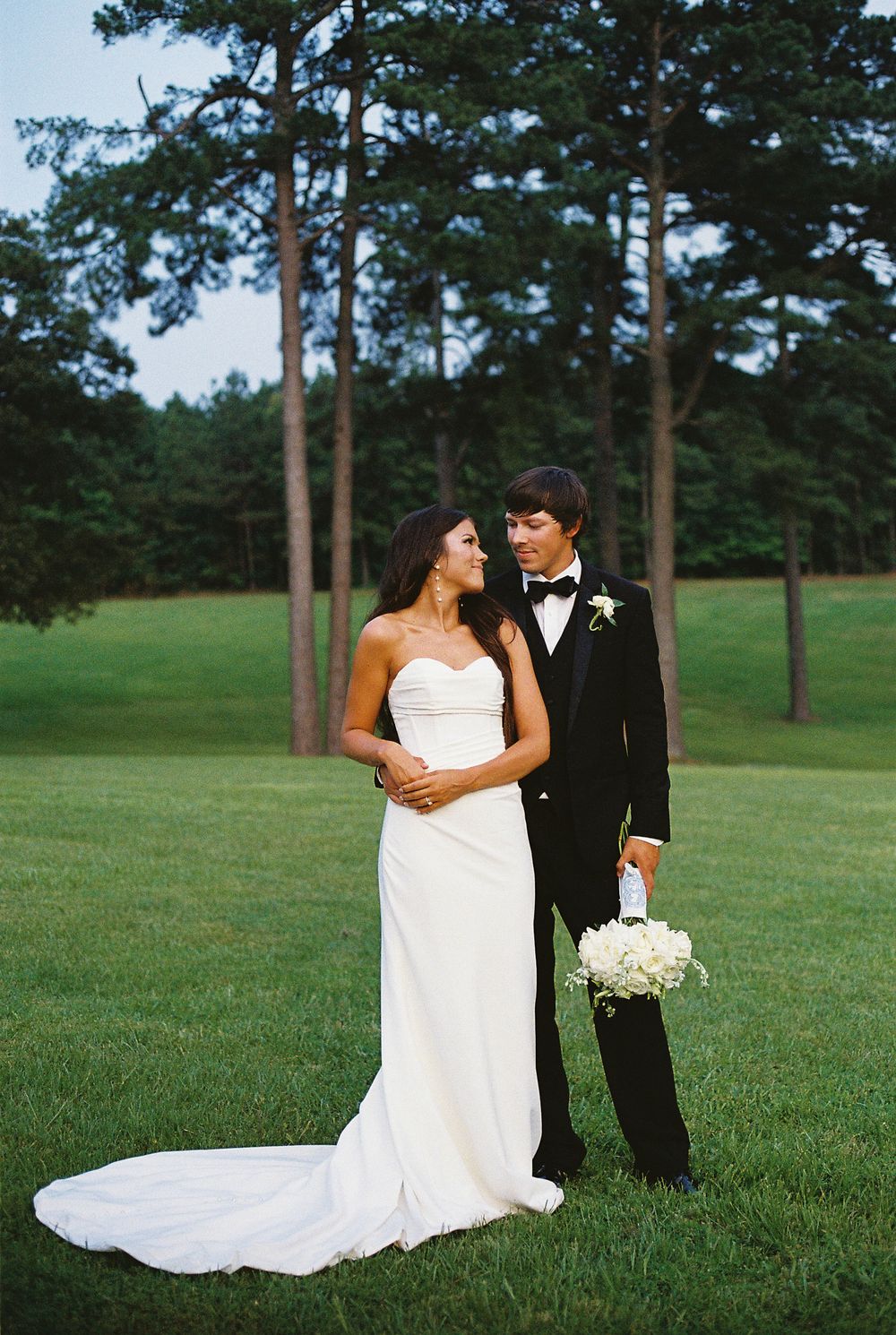Elegant wedding couple shares romantic moments in black tie attire against lush green golf course and tall pine trees.