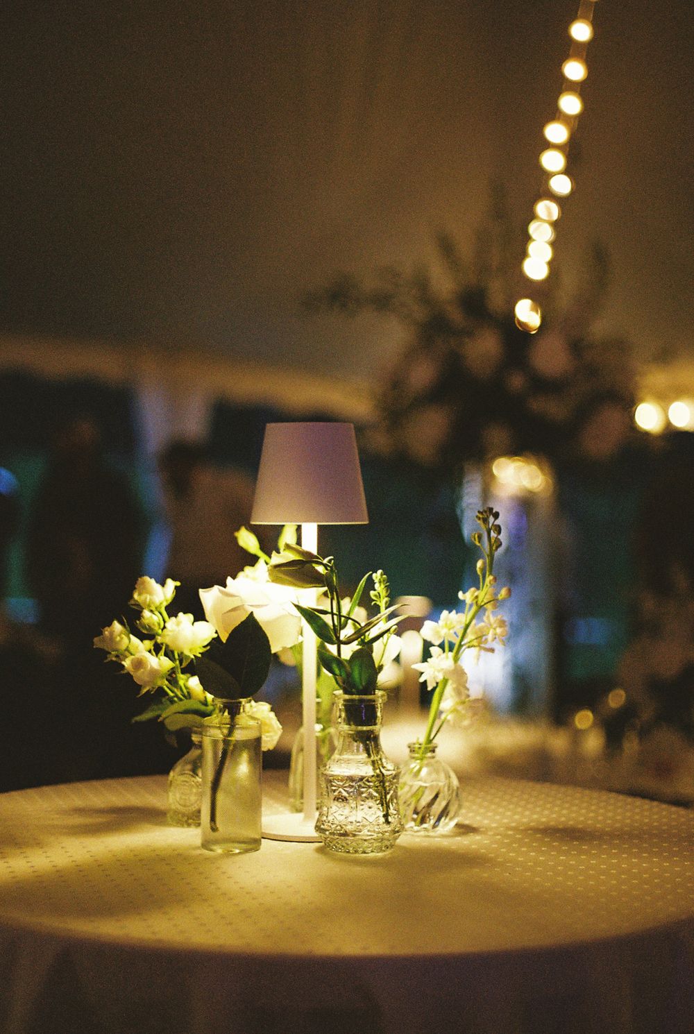 An elegant table centerpiece with white flowers in mason jars and string lights glowing in the background at a wedding reception.