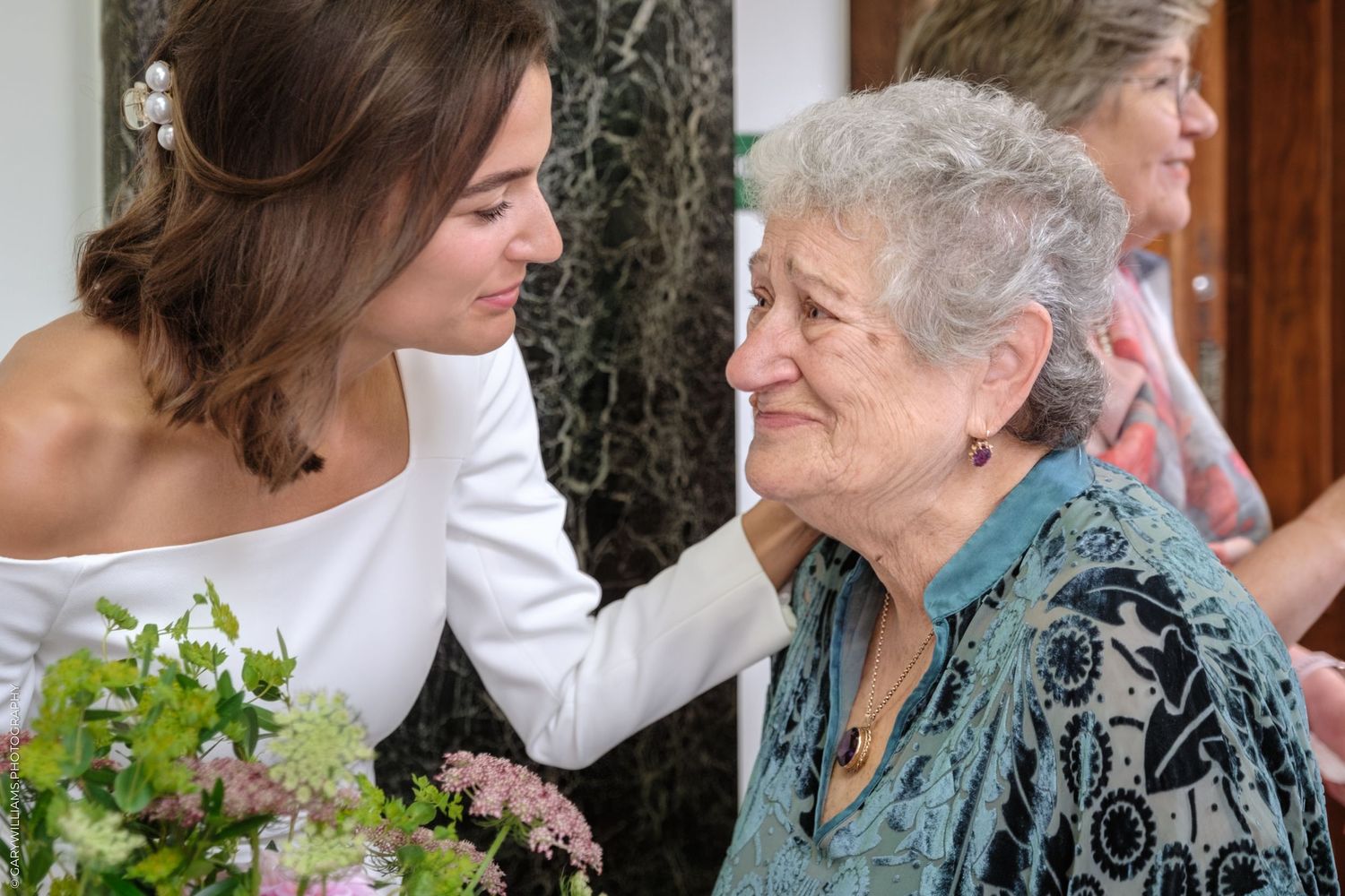 An elderly woman in teal floral attire and younger woman in white share a tender embrace during an outdoor celebration.