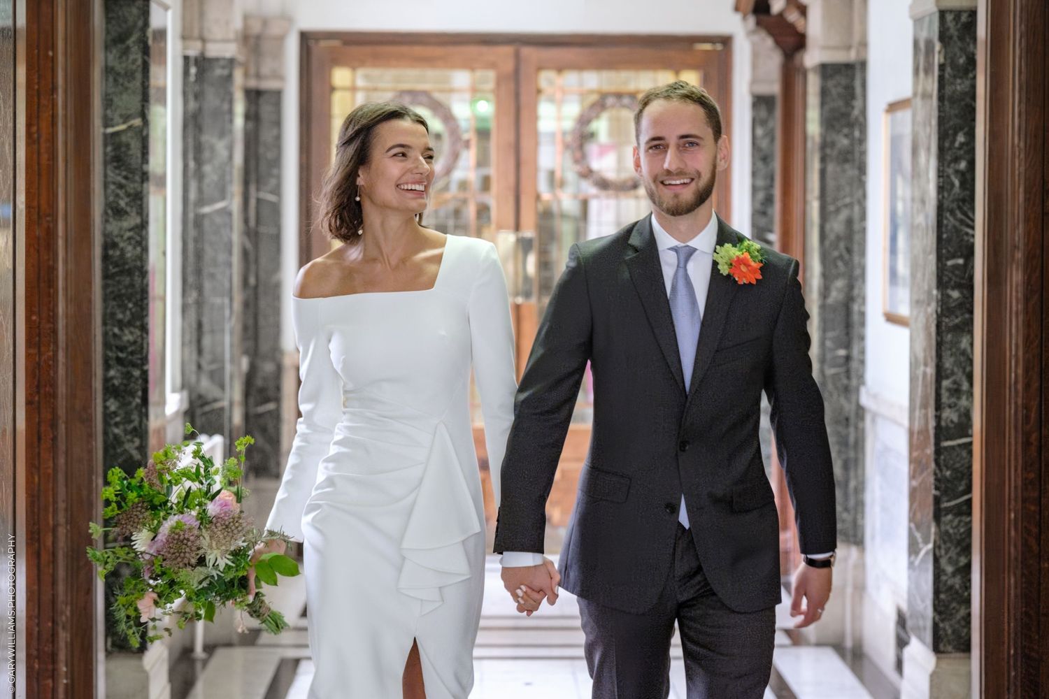 A couple in wedding attire walk together down an outdoor veranda surrounded by decorative columns and greenery.