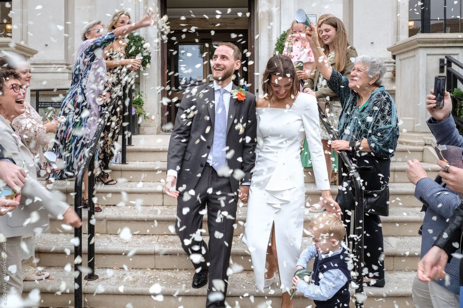 Joyful couple descending stone steps surrounded by wedding guests throwing confetti in celebration.