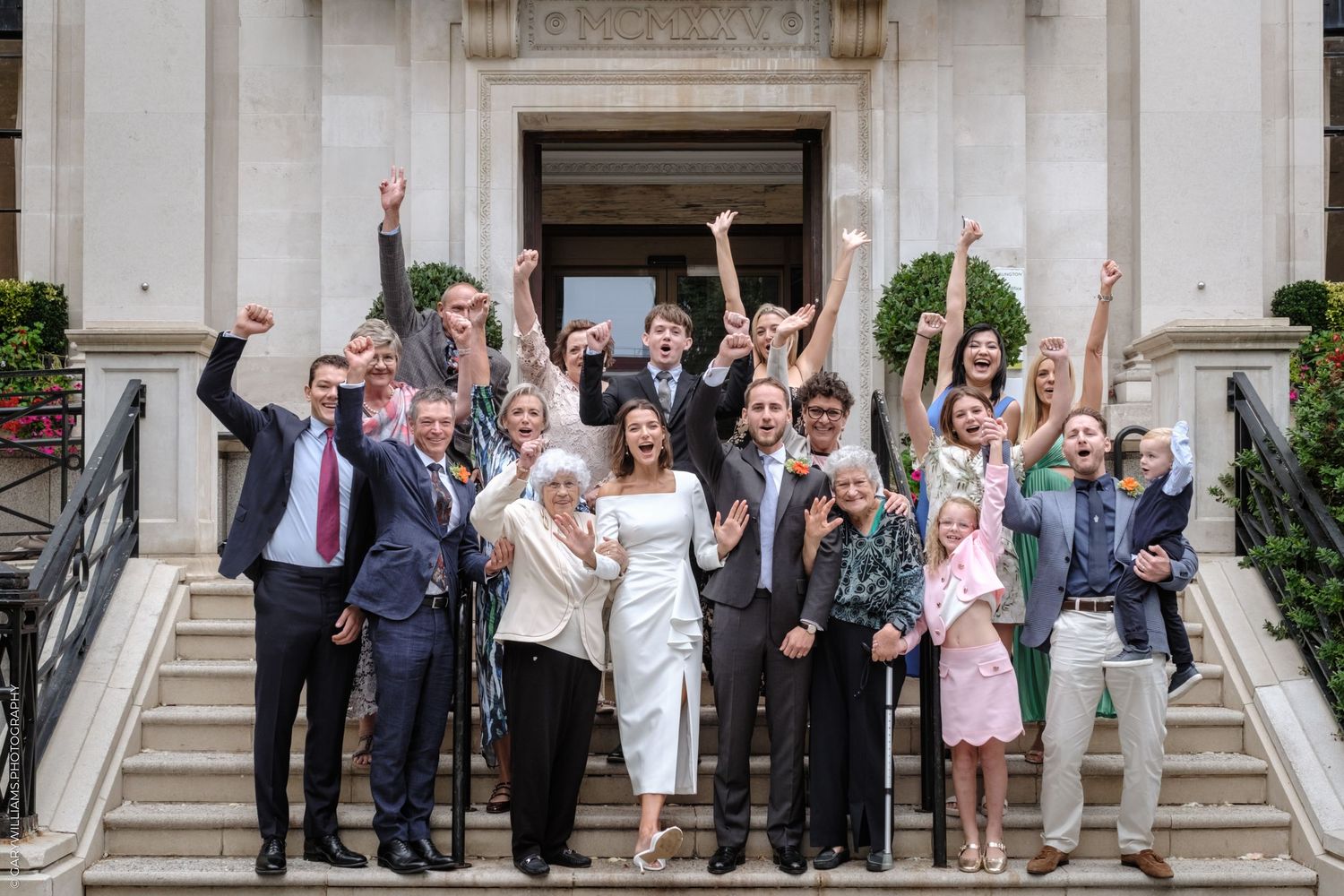 Wedding party members celebrate and wave while standing on grand stone steps of classic building.
