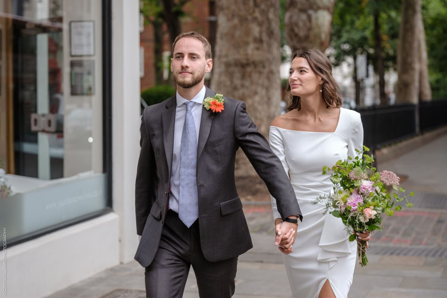 Wedding couple walks down sidewalk holding hands with greenery and boutiques in background.