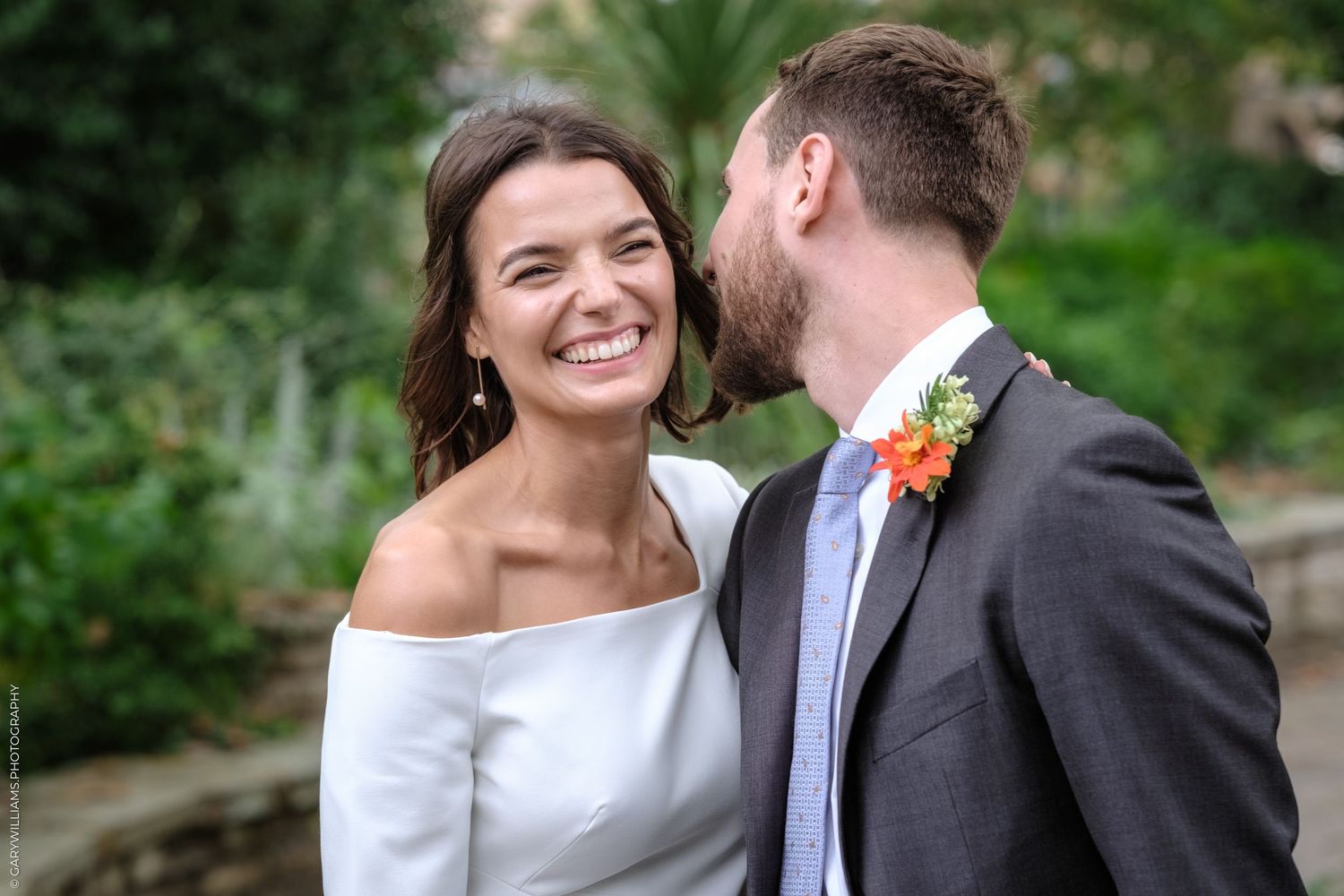 A happy couple shares a candid wedding moment outdoors, with the bride in white and groom in grey suit with orange boutonniere.