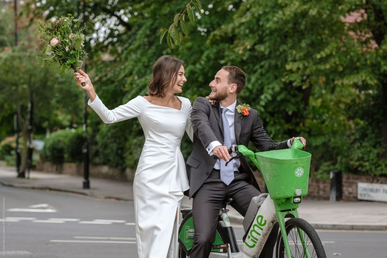 A couple joyfully celebrating their wedding day with a fun bike ride through a scenic park setting.