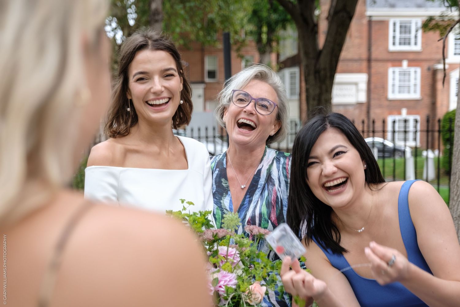 Three people share a joyful moment of laughter while holding colorful fresh flowers outdoors on a sunny day.