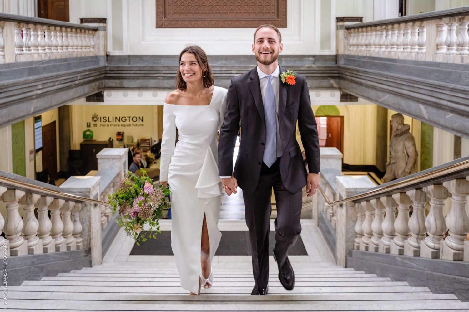 Two people in wedding attire walk down an ornate stone staircase together at a grand venue.