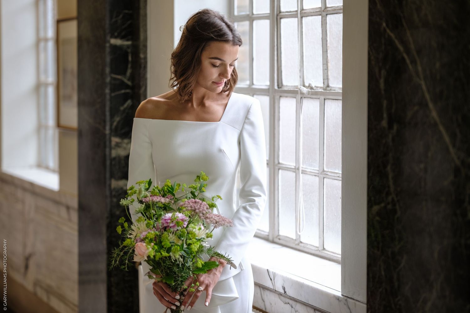 Someone in a flowy white dress stands beside interior windows holding a green bouquet in natural light.