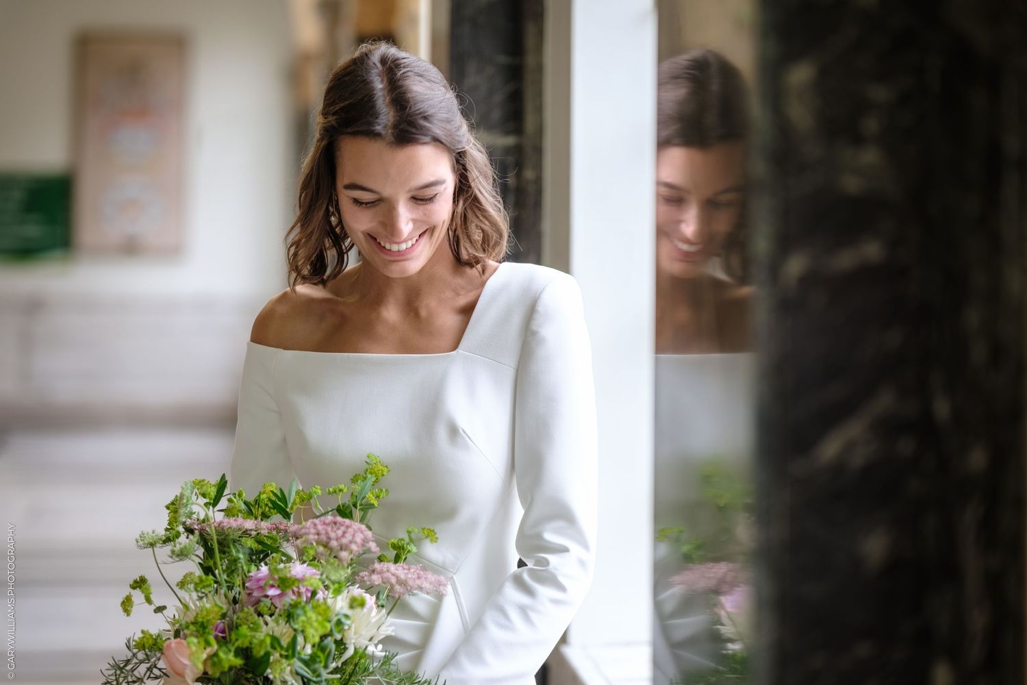 Bride in white dress walks gracefully down an elegant corridor with floral arrangements.