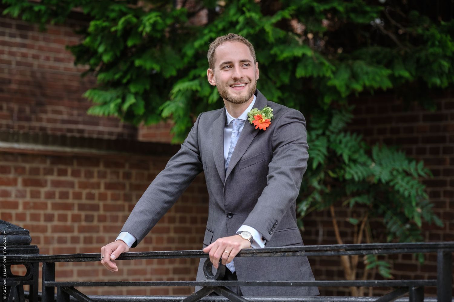 A groom in a gray suit with orange boutonniere poses against brick wall with green foliage backdrop.