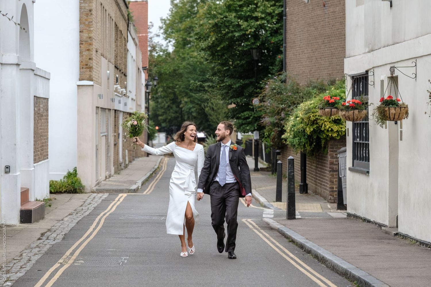 A couple walks joyfully down a charming tree-lined residential street in casual wedding attire.