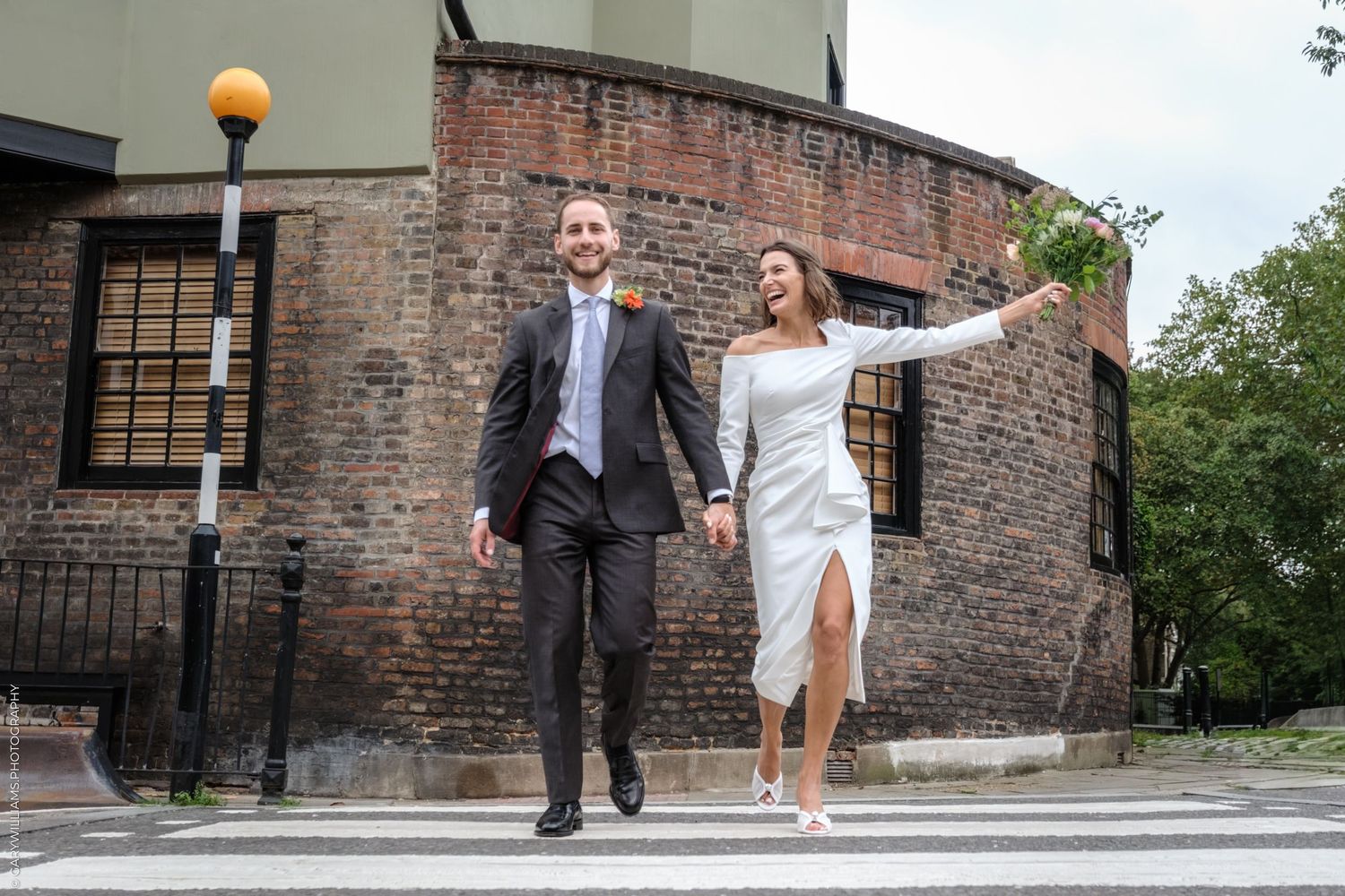 A couple in formal attire walks joyfully together on a sidewalk near a brick building with architectural details.