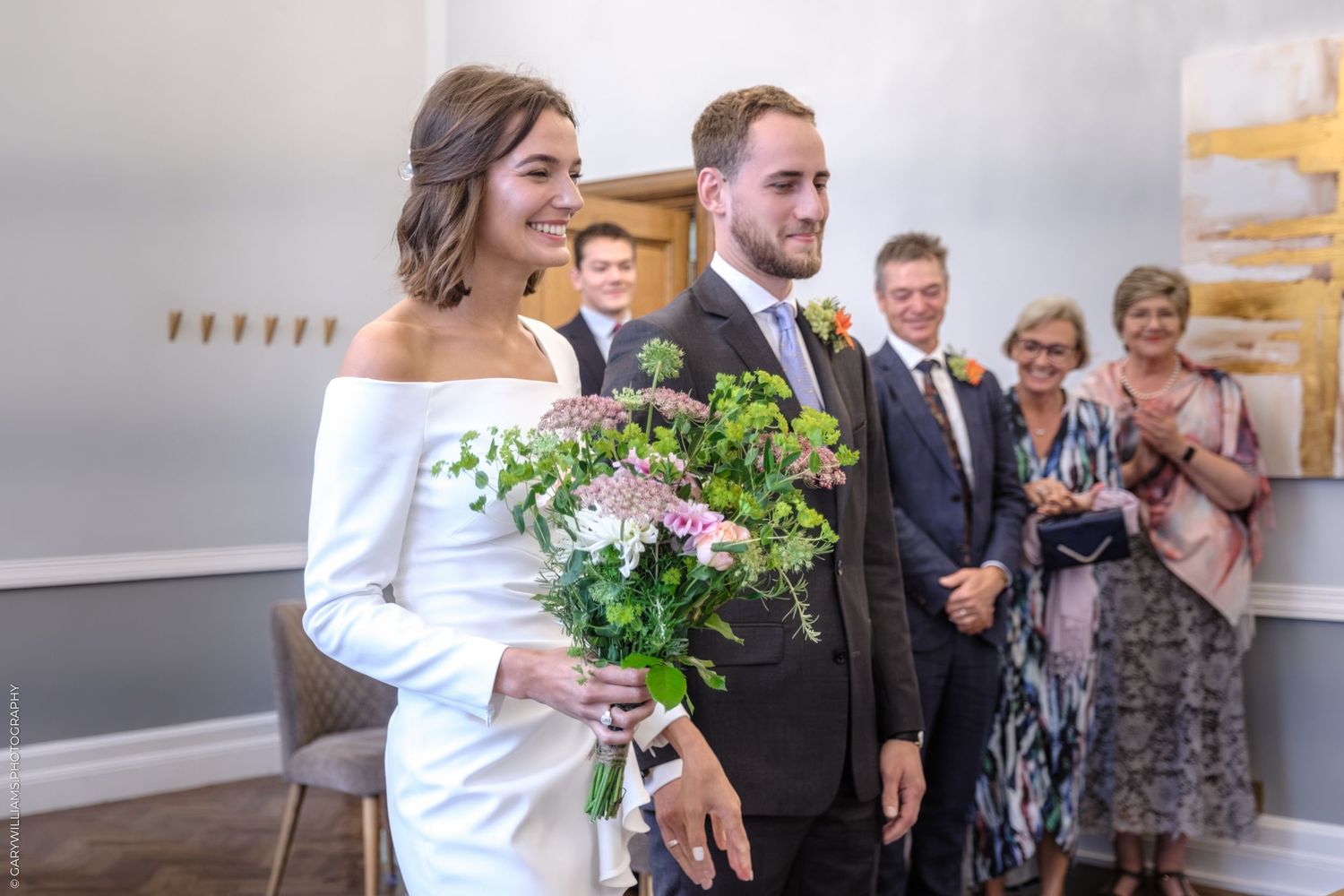 Bride in off-shoulder dress holds rustic bouquet while standing next to groom during wedding ceremony.