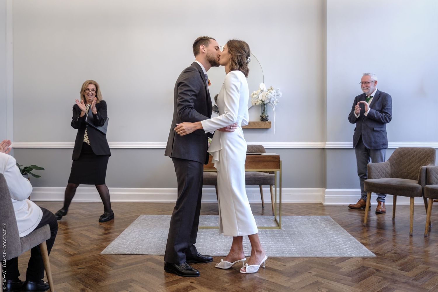 A couple shares a romantic embrace during an intimate wedding ceremony while guests look on and capture photos.