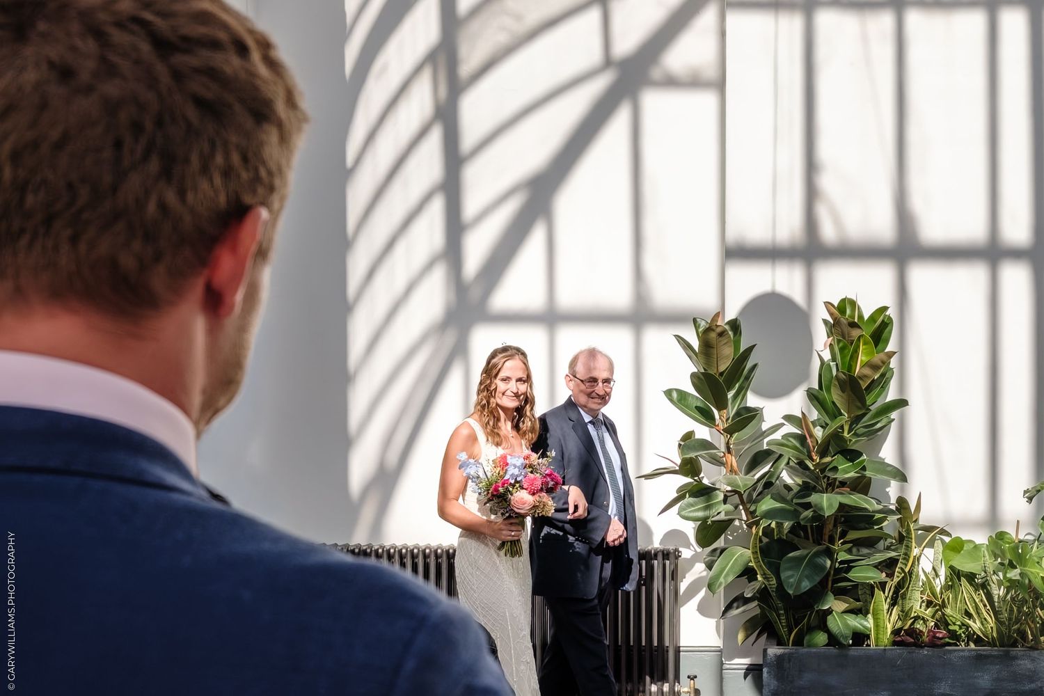 A wedding moment captured from behind a guest showing a happy couple entering through bright doors with white pillars.