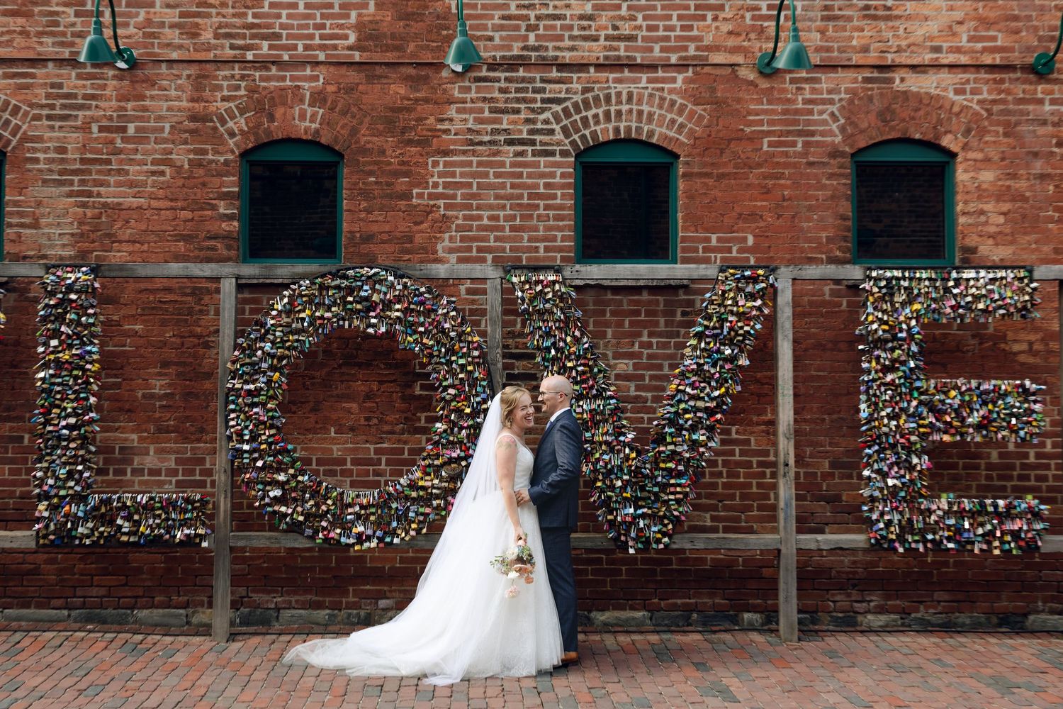 A couple poses in front of a rustic brick wall with floral 'LOVE' letters at a wedding venue.