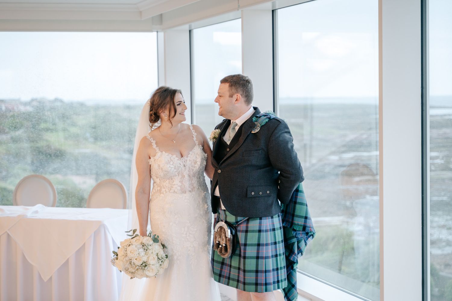 A couple poses by large windows, the groom in traditional Scottish wedding attire with a tartan kilt.