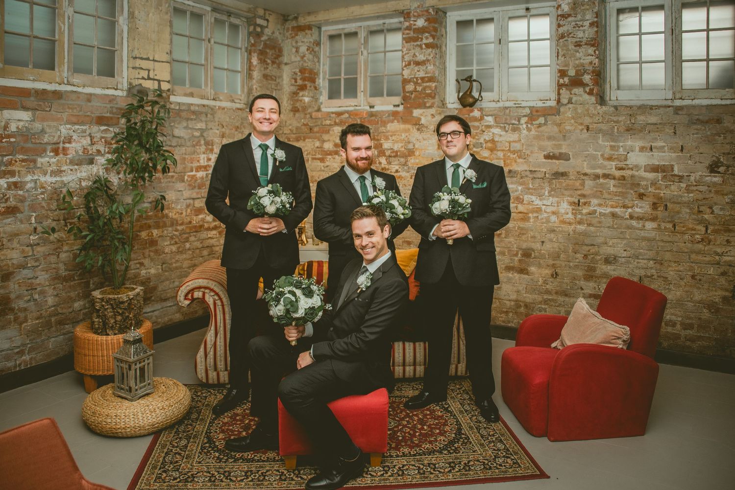 Wedding party poses against exposed brick wall with vintage red velvet chairs and potted plants in industrial venue.