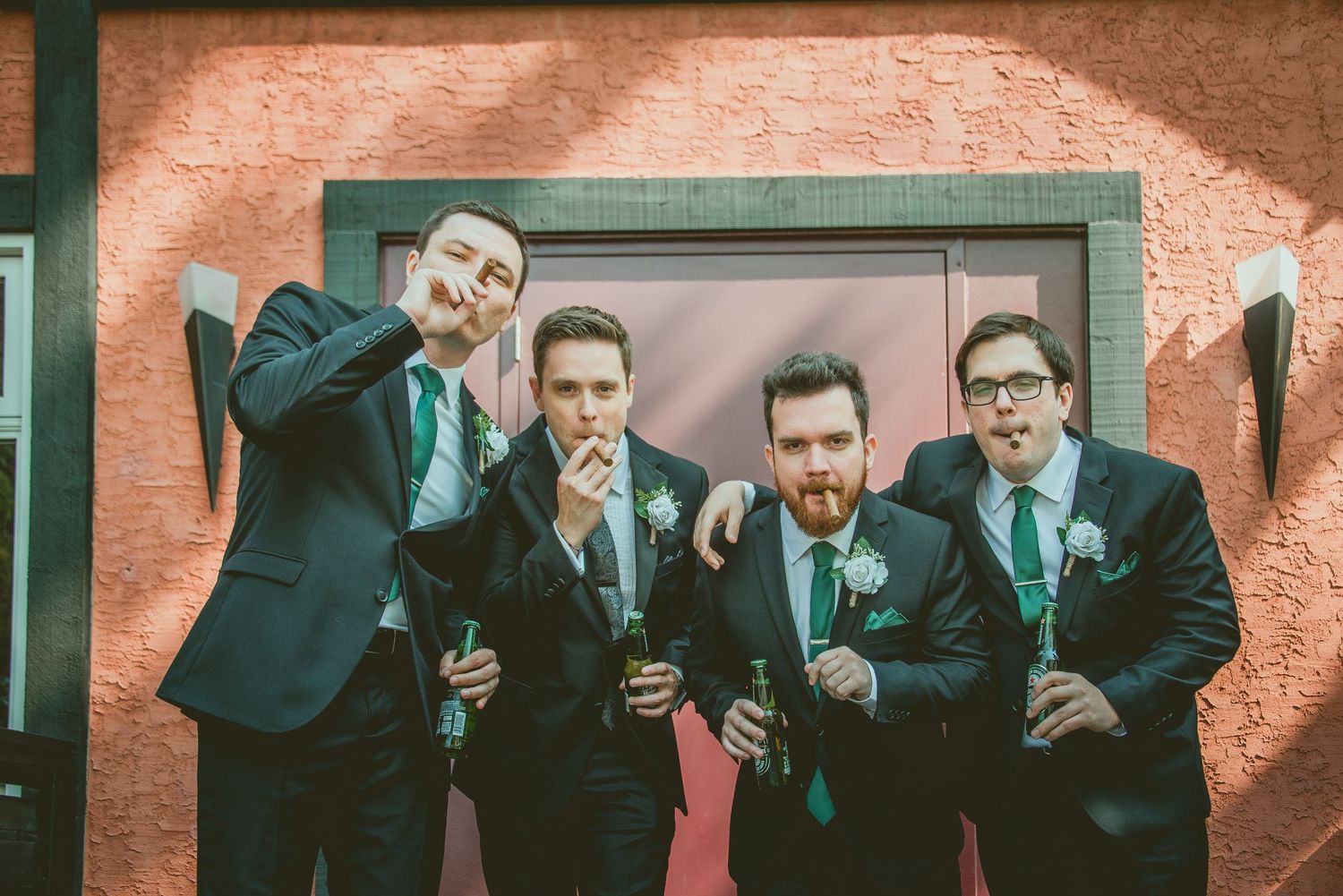 Four groomsmen in matching navy suits pose playfully against a coral-colored wall making funny gestures.