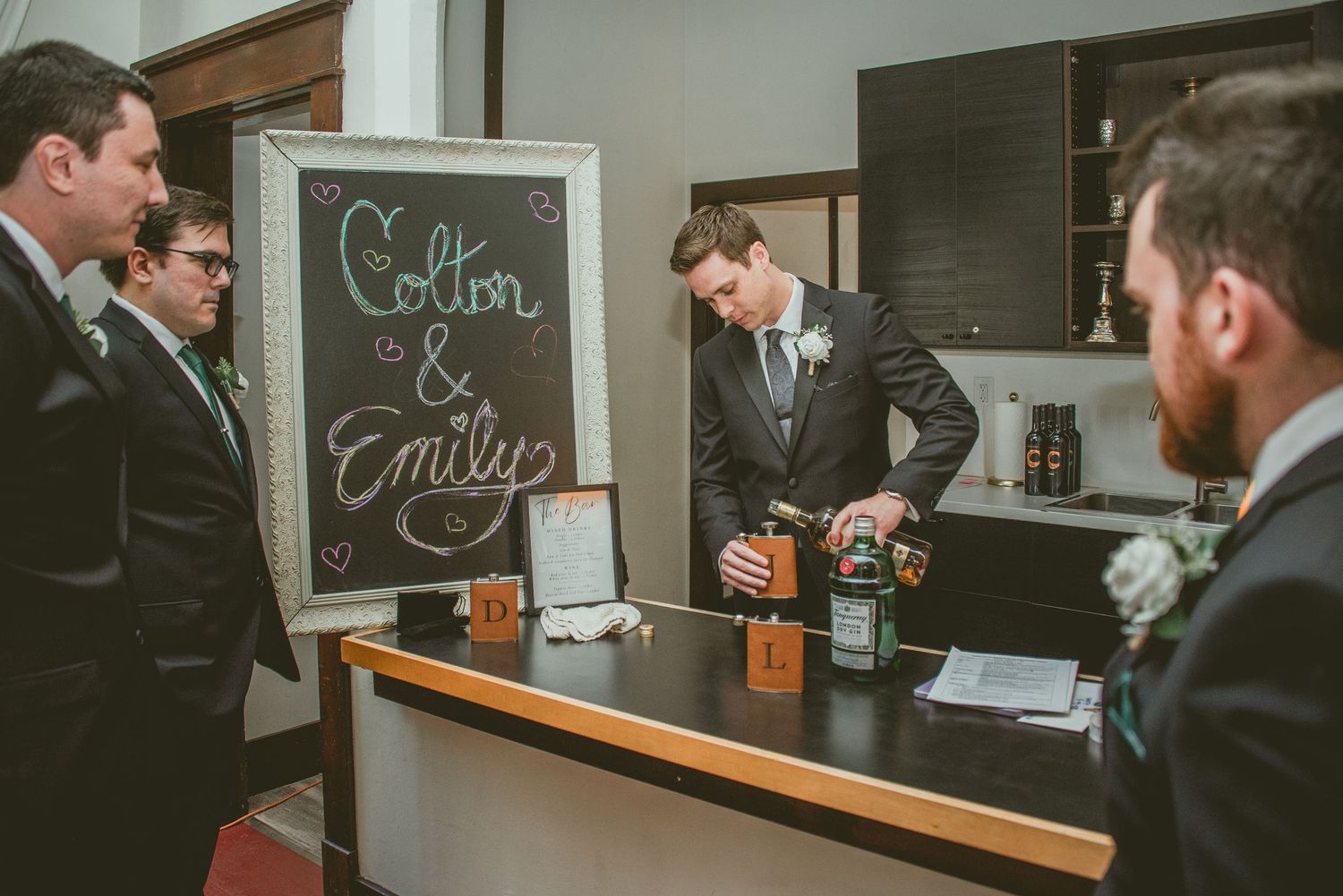 A group of people gather around a bar counter with chalkboard menus on the wall during what appears to be a tasting event.