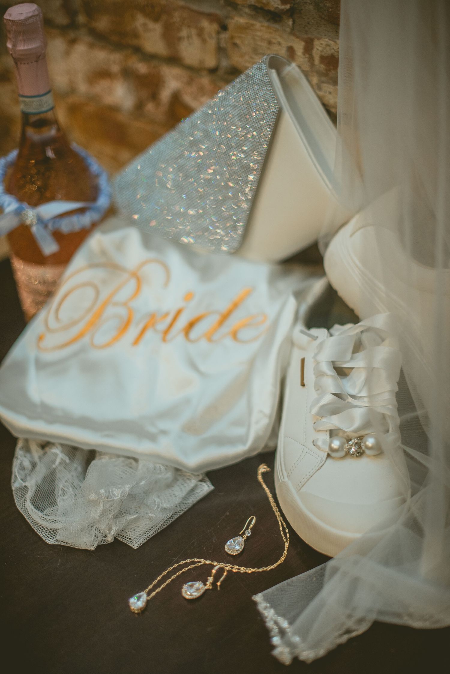 Wedding accessories including white sneakers, a sparkly heel, bride robe, and delicate pearl jewelry arranged on dark surface.