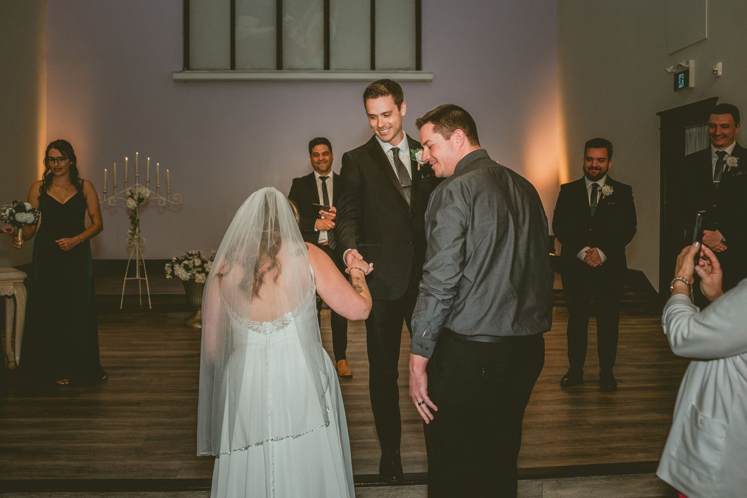 A wedding couple and their guests dance and celebrate in a dimly lit reception hall during an evening celebration.